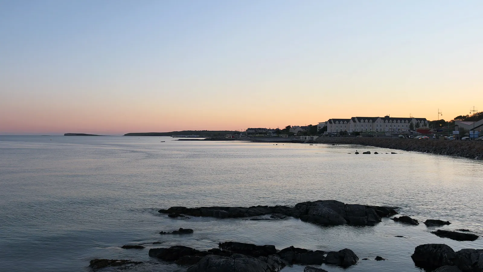 夕暮れの穏やかなゴールウェイ湾に浮かぶ黒い岩々、その向こう岸にはソルトヒルの海岸通りに灯りがともる建物が連なっています