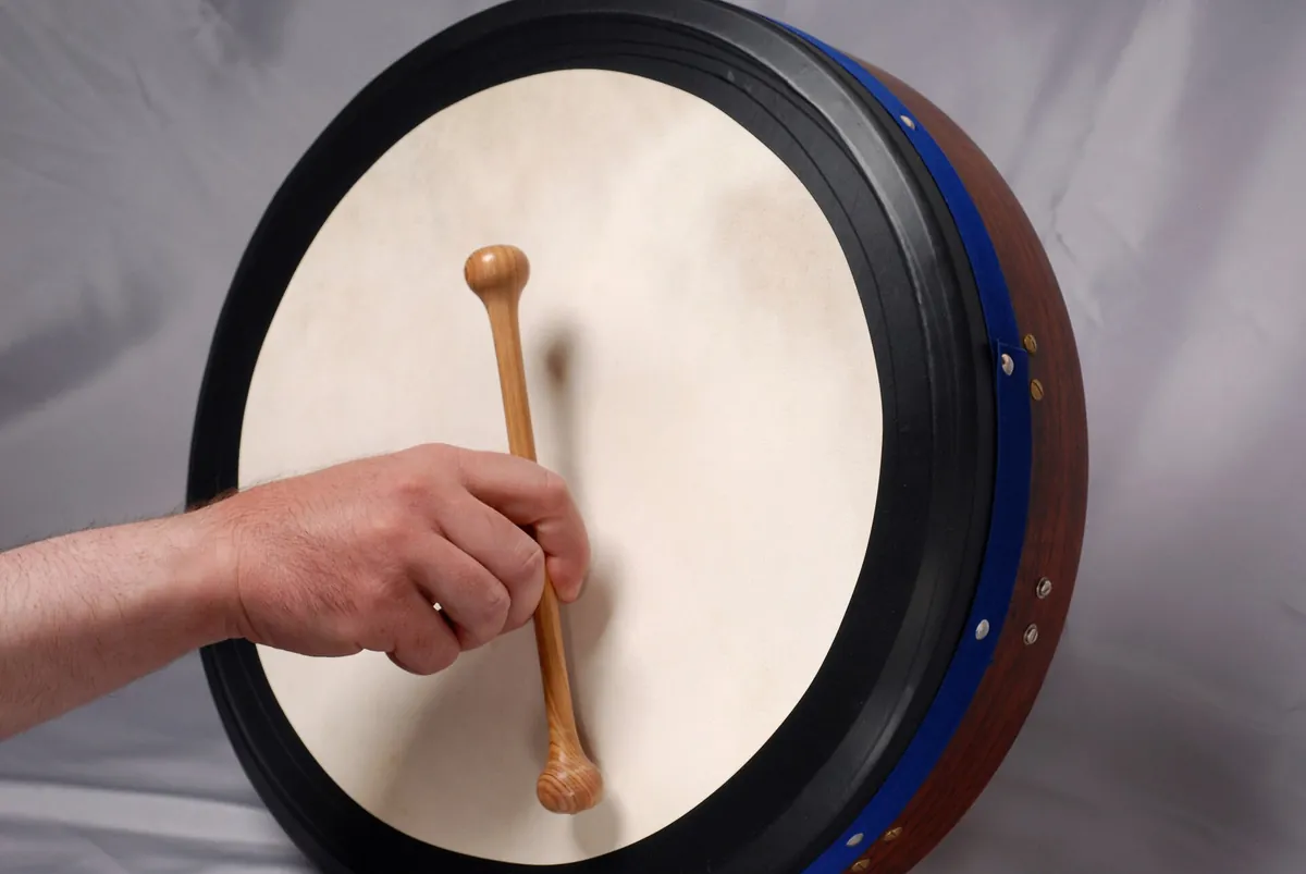 Un bodhrán con un parche de piel de cabra clara y un borde azul, una mano sosteniendo una baqueta de madera contra la piel
