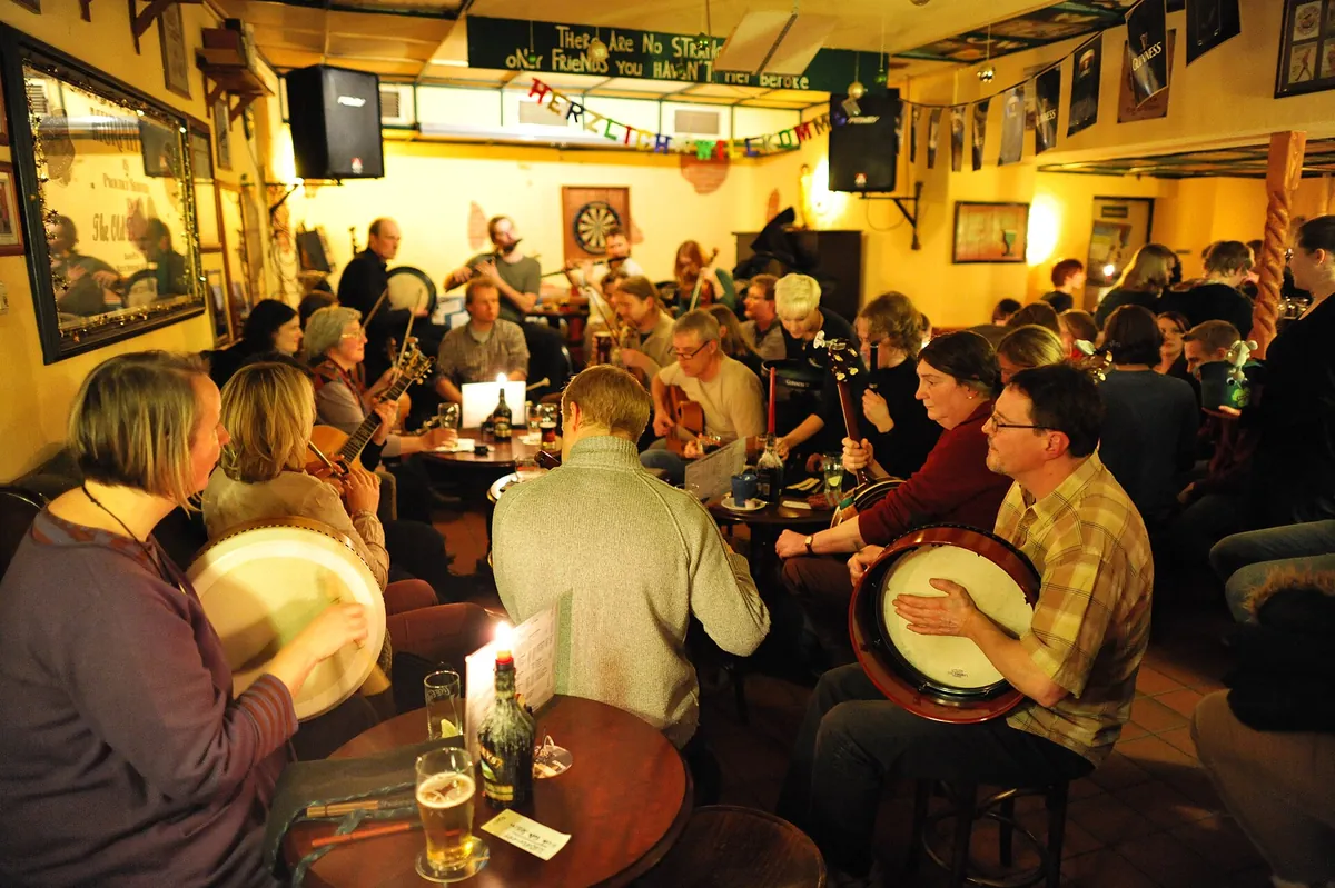 Músicos tocando violines y guitarras en una mesa de un pub, con vasos y una barra de madera al fondo