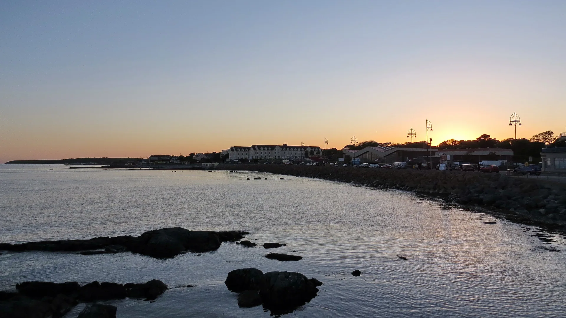 El paseo marítimo de Salthill al atardecer, con las farolas del paseo y edificios bajos recortados tras un malecón de piedra y las tranquilas aguas de la bahía de Galway
